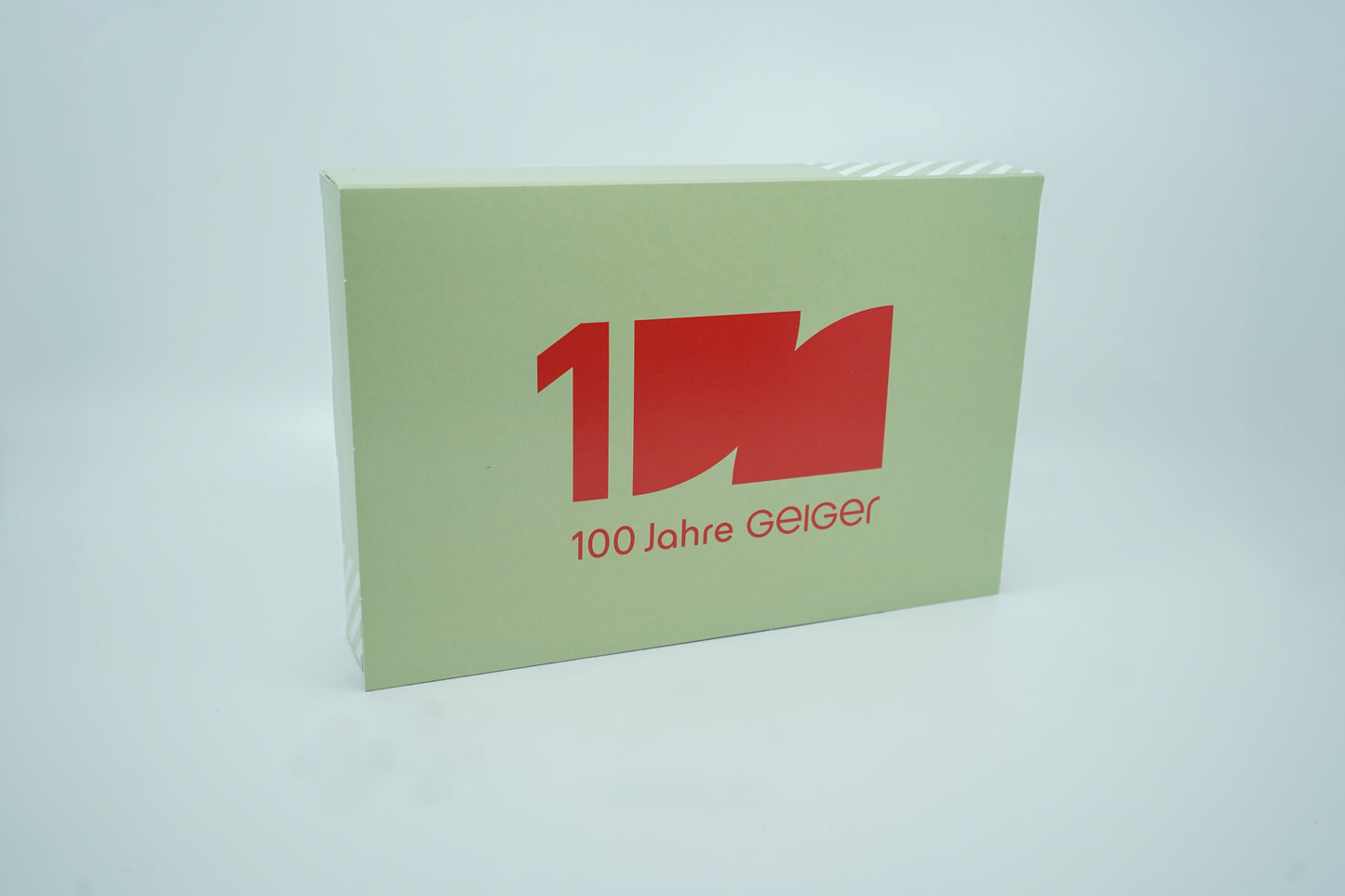 Miniaturmodelle zweier MAN Geiger-LKW mit Anhänger (Oldtimer und Fantasiemodell) in Geiger-Grün und rotem "Geiger" Schriftzug im "100 Jahre Geiger" Klappkarton. Auf dem Klappkarton ist der Schriftzug 100 Jahre Geiger abgebildet.