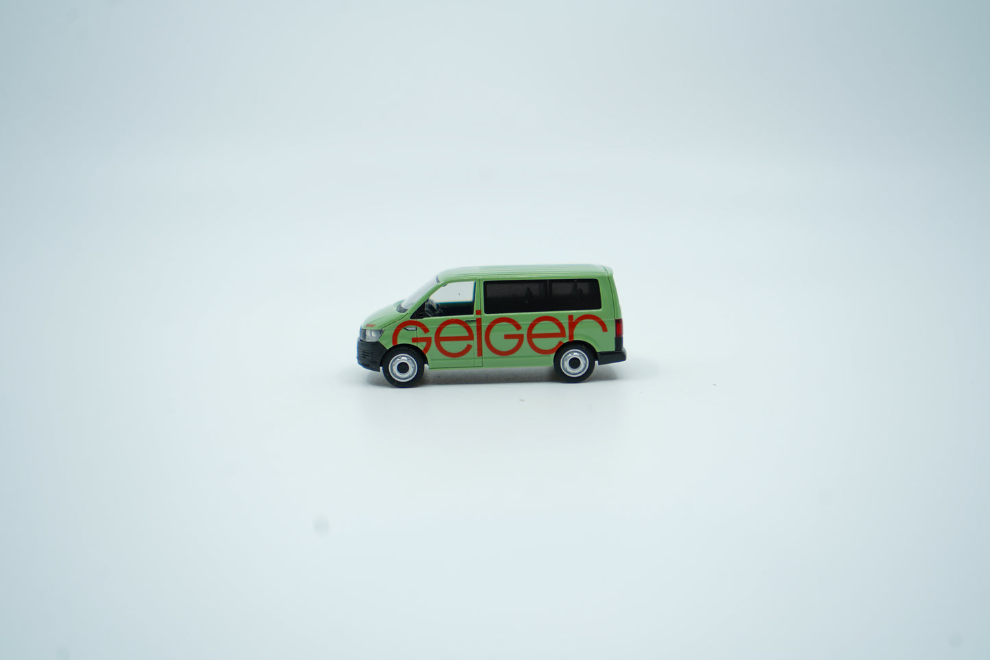 Miniaturmodell eines Geiger VW-Bus in Geiger-Grün mit rotem „Geiger“-Schriftzug, Verpackung inklusive.