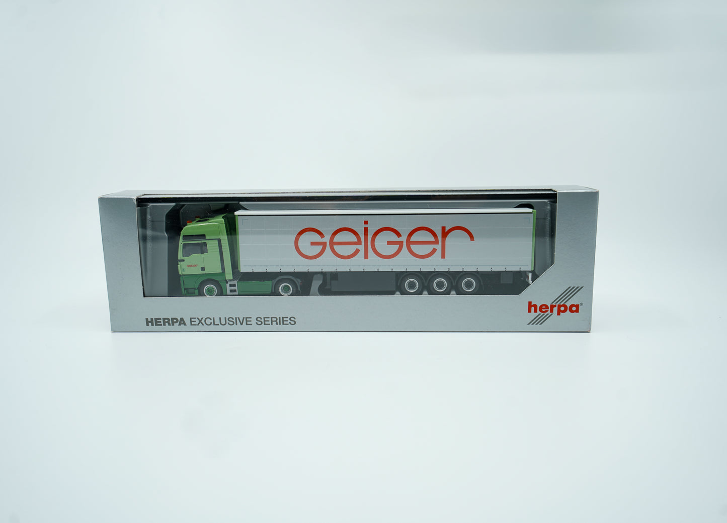 Miniaturmodell eines MAN Geiger-LKW in Geiger-Grün mit grauem "Gardinenplanen" Anhänger mit rotem "Geiger" Schriftzug. Verpackung inklusive.