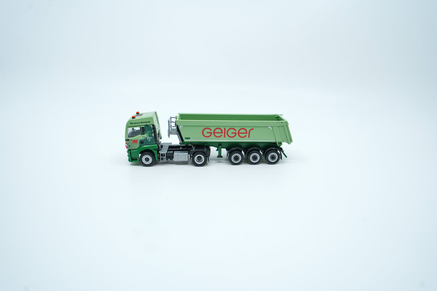 Miniaturmodelle zweier MAN Geiger-LKW mit Anhänger (Oldtimer und aktuelles Modell) in Geiger-Grün und rotem "Geiger" Schriftzug mit 100 Jahre Gestaltung im "100 Jahre Geiger" Klappkarton. Auf dem Klappkarton ist Wilhelm Geiger, der Firmengründer abgebildet.