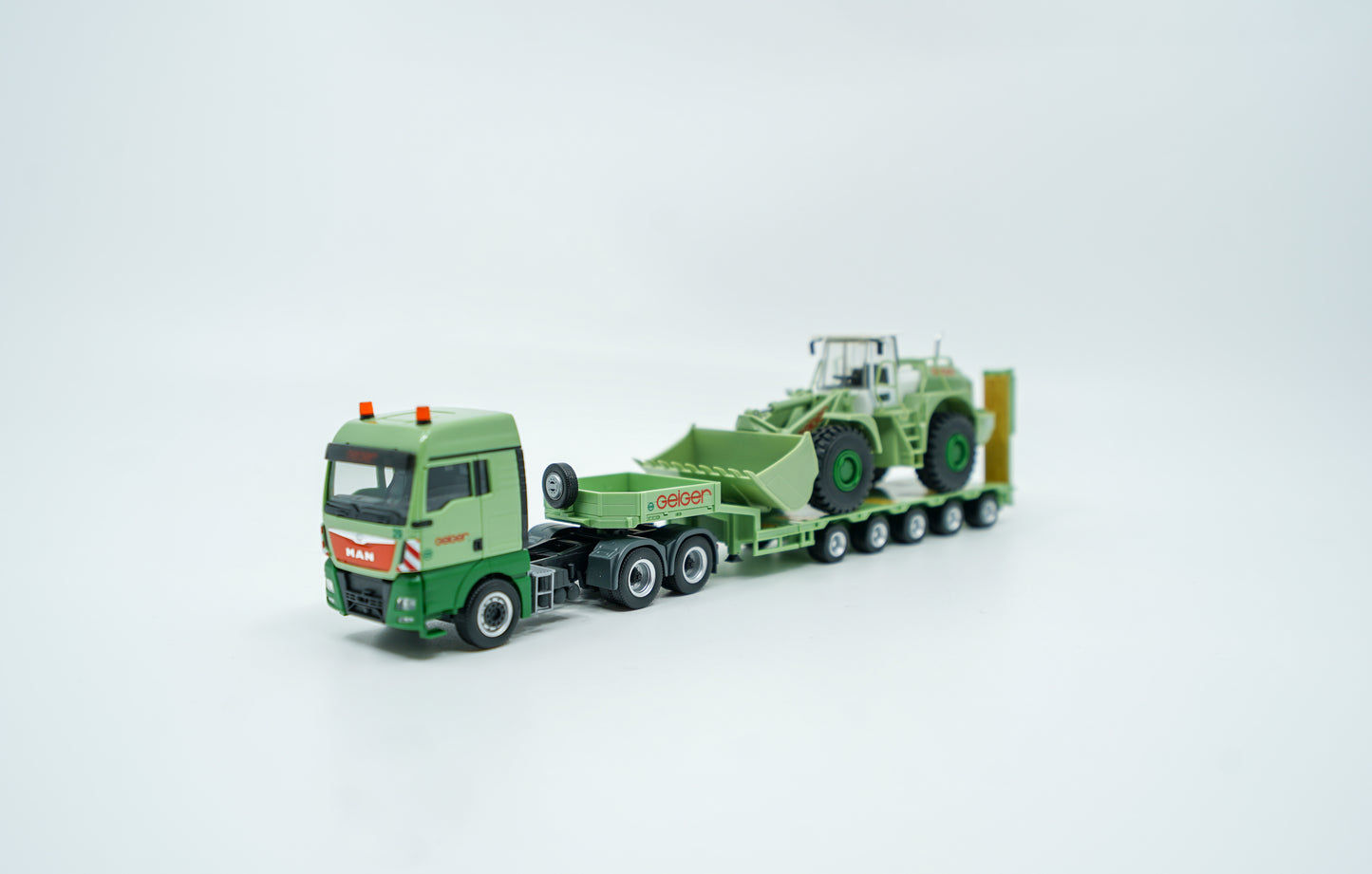 Miniaturmodell eines MAN Geiger-Tiefladers mit Radlader in Geiger-Grün und roten "Geiger" Schriftzügen. Verpackung inklusive.