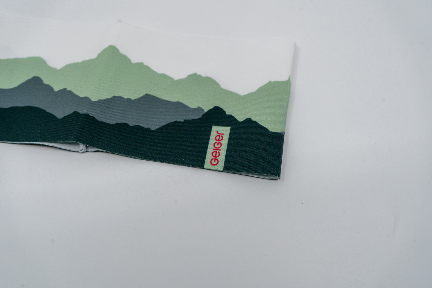 Sportstirnband mit verschiedenen Bergen abgebildet in mehreren Grüntönen und "Geiger" Schriftzug auf einem Patch.