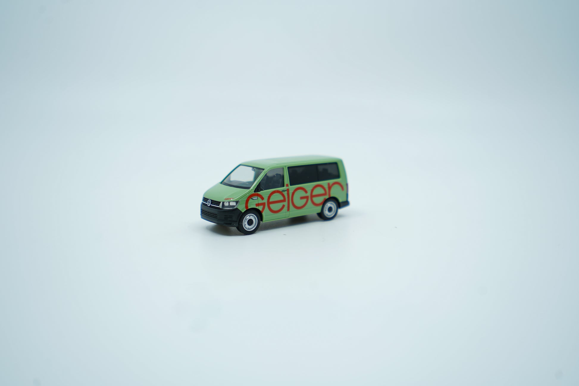 Miniaturmodell eines Geiger VW-Bus in Geiger-Grün mit rotem „Geiger“-Schriftzug, Verpackung inklusive.
