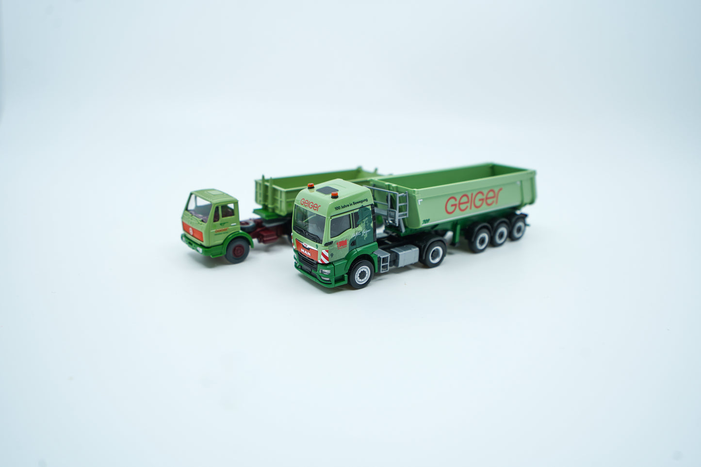 Miniaturmodelle zweier MAN Geiger-LKW mit Anhänger (Oldtimer und aktuelles Modell) in Geiger-Grün und rotem "Geiger" Schriftzug mit 100 Jahre Gestaltung im "100 Jahre Geiger" Klappkarton. Auf dem Klappkarton ist Wilhelm Geiger, der Firmengründer abgebildet.