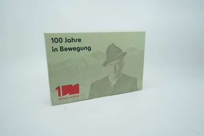 Miniaturmodelle zweier MAN Geiger-LKW mit Anhänger (Oldtimer und aktuelles Modell) in Geiger-Grün und rotem "Geiger" Schriftzug mit 100 Jahre Gestaltung im "100 Jahre Geiger" Klappkarton. Auf dem Klappkarton ist Wilhelm Geiger, der Firmengründer abgebildet.