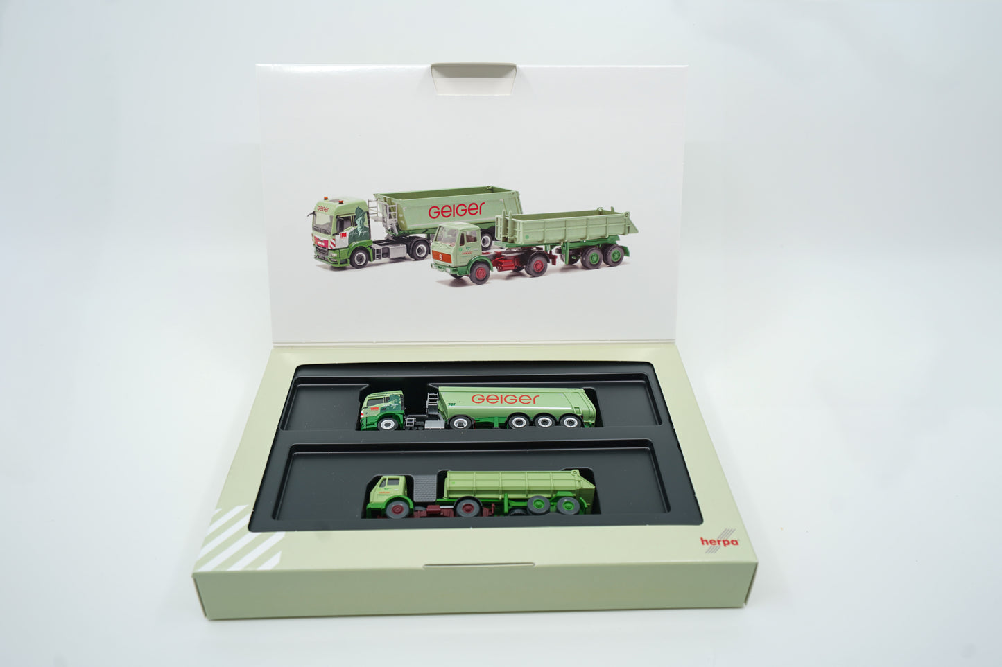 Miniaturmodelle zweier MAN Geiger-LKW mit Anhänger (Oldtimer und aktuelles Modell) in Geiger-Grün und rotem "Geiger" Schriftzug mit 100 Jahre Gestaltung im "100 Jahre Geiger" Klappkarton. Auf dem Klappkarton ist Wilhelm Geiger, der Firmengründer abgebildet.